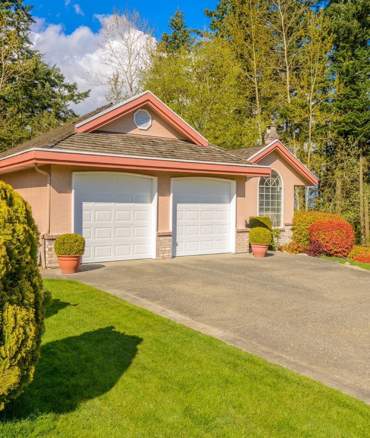 Front view of a peach-colored, two-car garage home with white doors, pink trim, and manicured shrubs in a sunny yard.