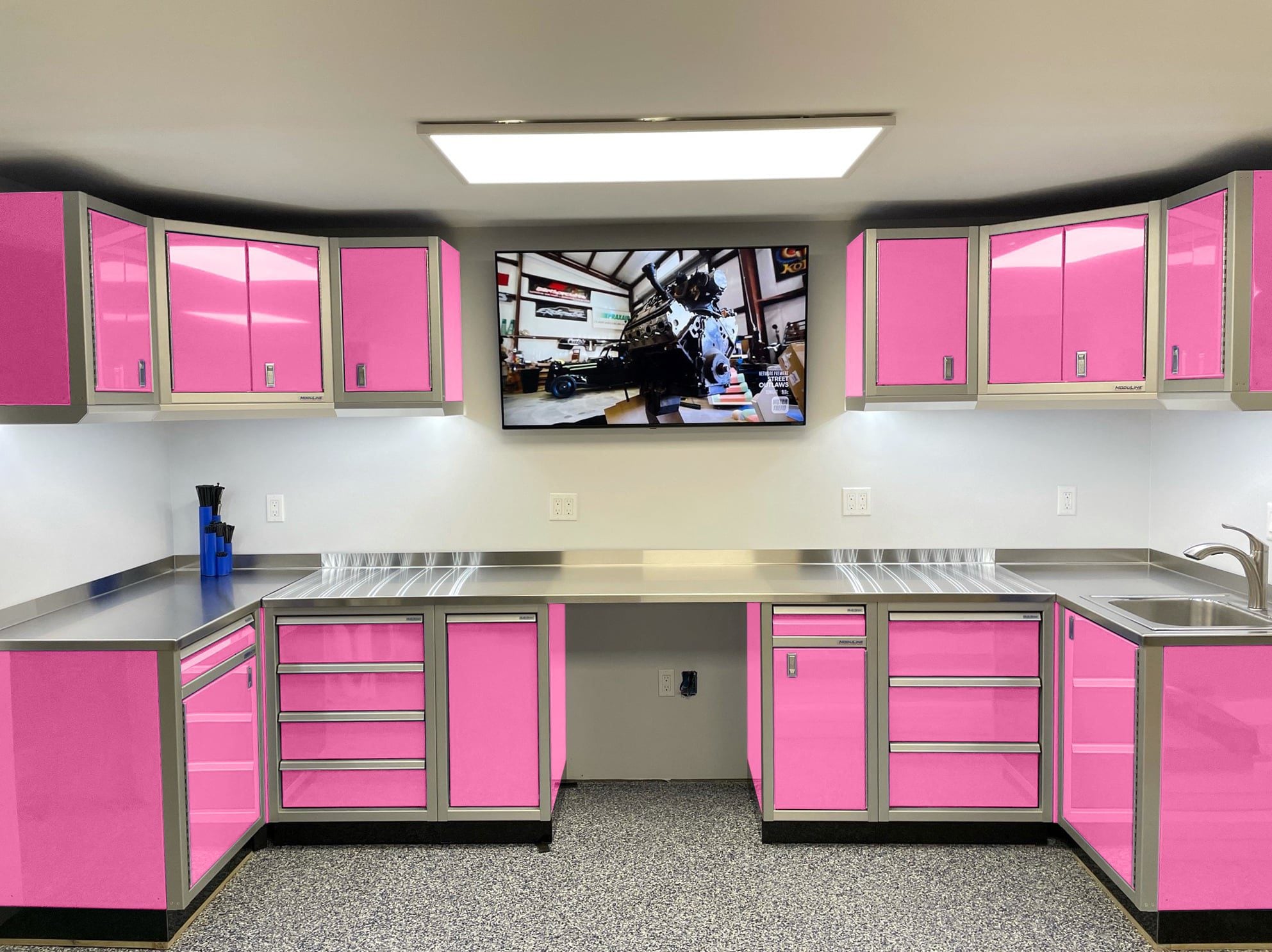 Pink and gray automotive workshop with pink wall and base cabinets, stainless countertops, and a wall-mounted monitor.