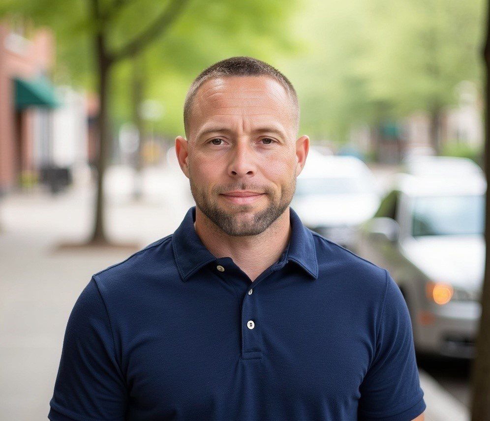 Portrait of a man with short hair in a navy polo standing on a tree-lined urban sidewalk, looking at the camera.