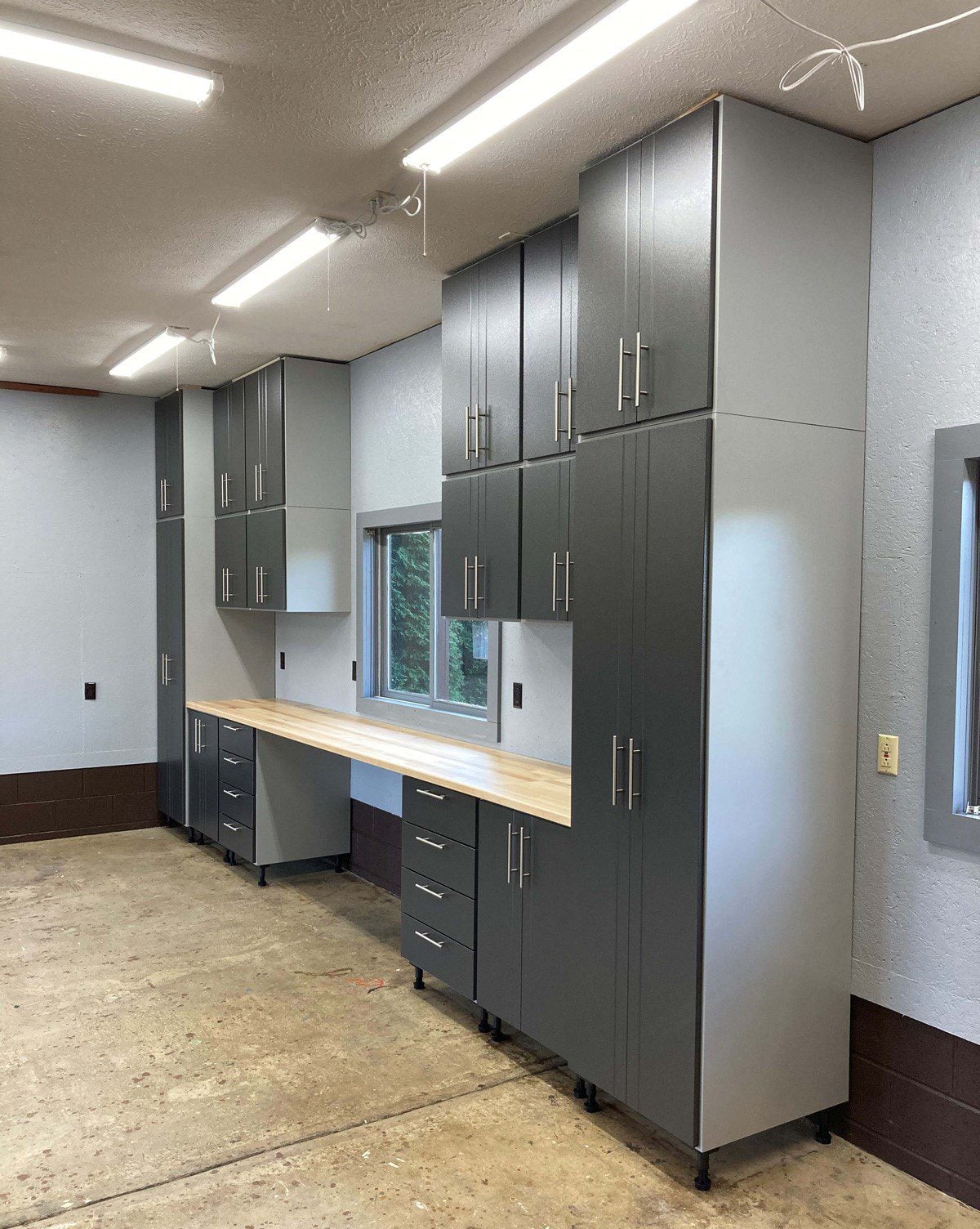 Workshop storage area with tall gray cabinets and a light wood countertop running along the wall beneath a window.