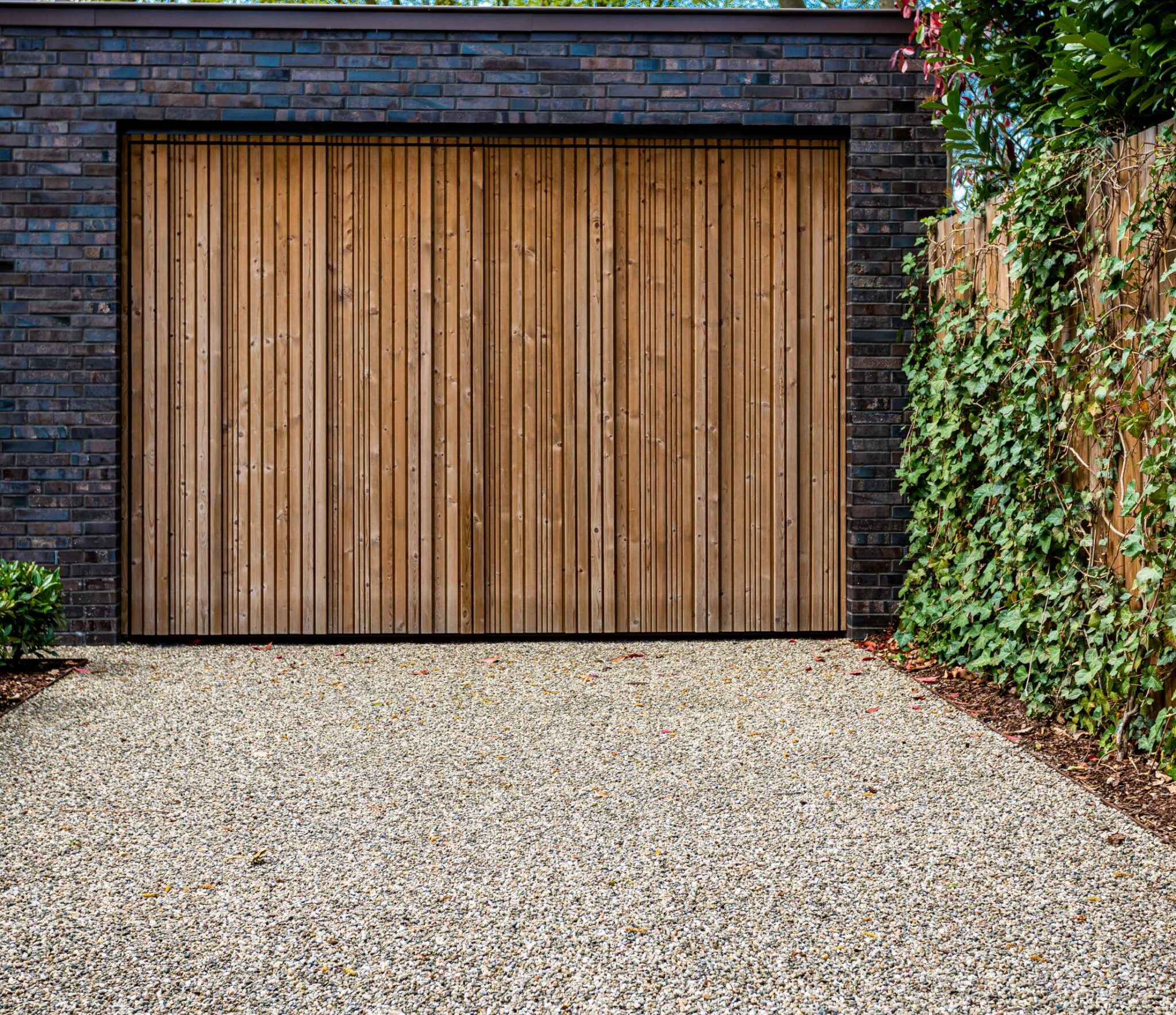 Brown wooden vertical-slatted garage door set in a dark brick facade, with ivy along the right and a gravel driveway in front.