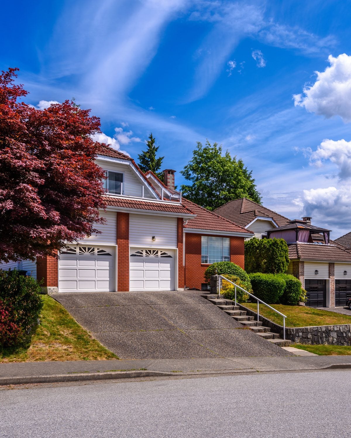 Front view of a two-car garage home with brick columns, white siding, and a red-tiled roof, a red-leaved tree on the left and a blue sky above.