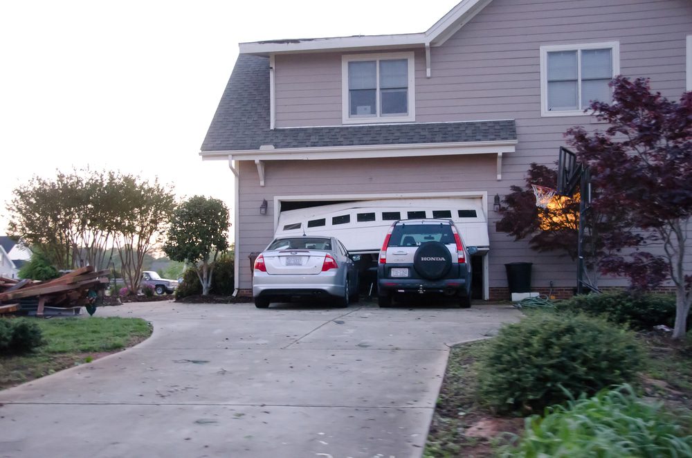 Two cars parked in a concrete driveway in front of a beige two-story house with a closed double garage door and a basketball hoop on the right.
