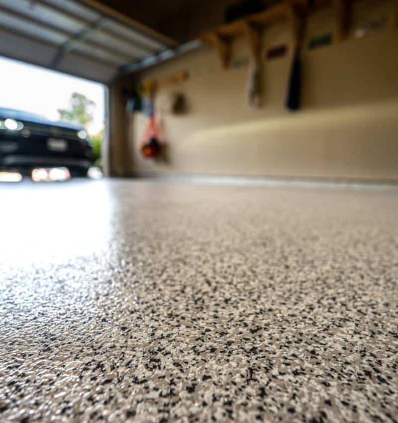 Low-angle view of a speckled epoxy garage floor with a blurred car parked in an open garage in the background.