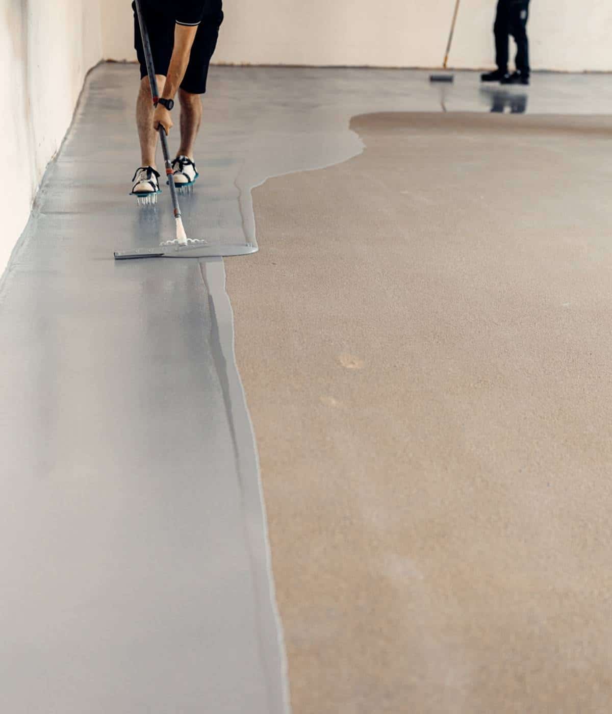 Worker in shorts and sneakers smooths a wet concrete surface with a trowel near a wall in an indoor space.