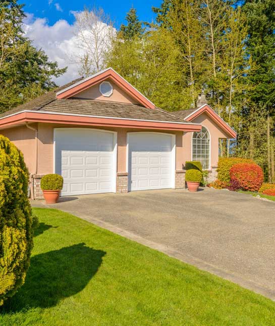 Two-car garage with white doors on a pink house, front yard with potted shrubs and a green lawn.”,