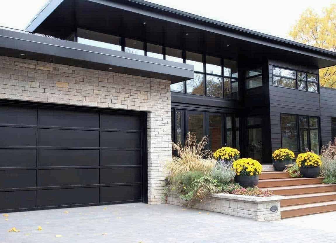 Modern house entrance with a large black sectional garage door, stone walls, and wooden steps lined with yellow chrysanthemums in pots on the right side.